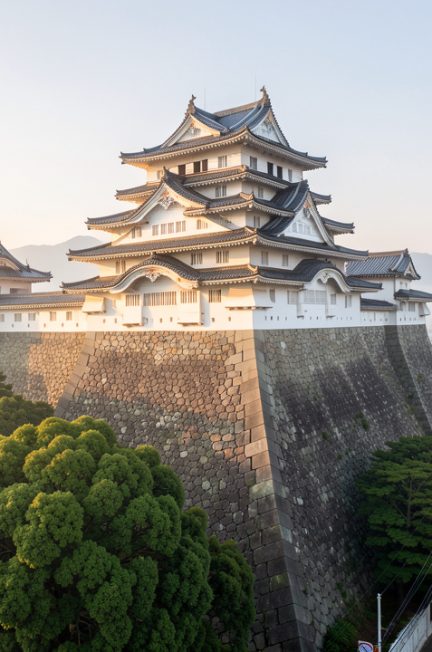 Vue immersive du château de Kumamoto au petit matin, avec ses remparts en pierre, une végétation luxuriante et les rues calmes de la ville japonaise.