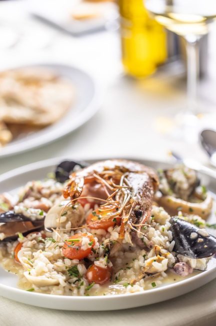 Une poêle de paella garnie de fruits de mer et de légumes, accompagnée d’un verre de vin blanc sur une table.