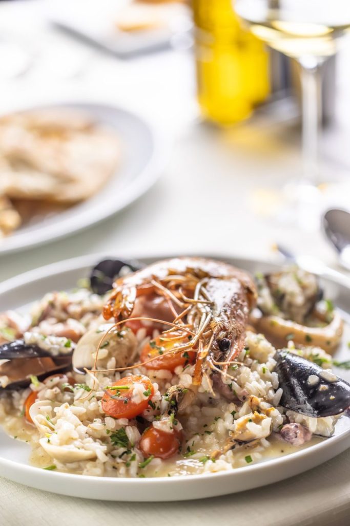 Une poêle de paella garnie de fruits de mer et de légumes, accompagnée d’un verre de vin blanc sur une table.